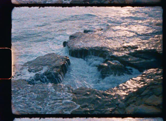 Super 8mm film captures slow-motion waves crashing on a rocky coast, followed by a beautiful sunset view over the ocean with a distant island