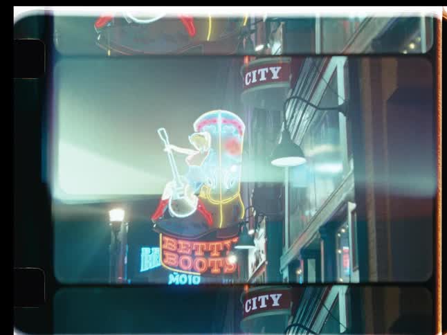 A vintage film-style zoom out reveals a vibrant neon sign of a cowgirl sitting on a large boot on a city street at night