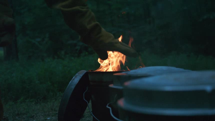 A person in a protective glove warms their hand over a fire in a metal can in the woods in slow motion