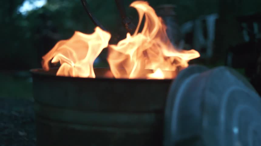 Mesmerizing slow-motion flames dance and flicker inside a rustic metal barrel during an evening bonfire