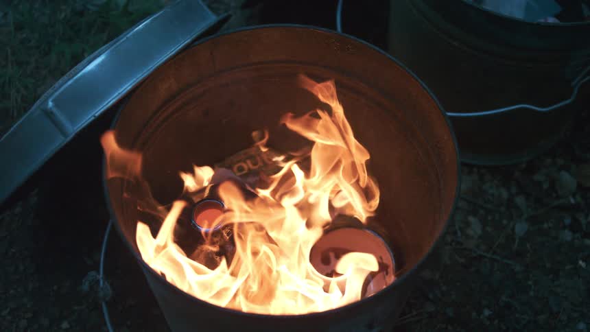 Slow motion footage shows bright orange flames consuming objects inside a metal burn barrel