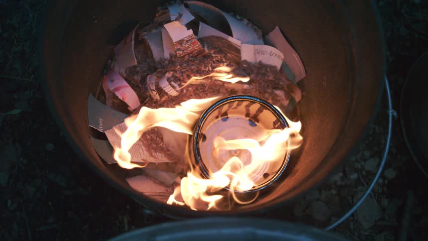A mesmerizing top-down, slow-motion view of a ceramic bowl being pit fired in a metal bucket, surrounded by burning paper and combustible materials