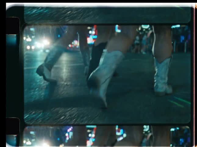 Vintage film captures a low-angle view of women in cowboy boots walking down a busy city street at night surrounded by neon lights