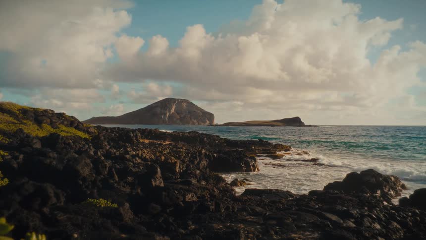Gentle ocean waves crash against a rugged volcanic rock shoreline with a tropical island in the distance