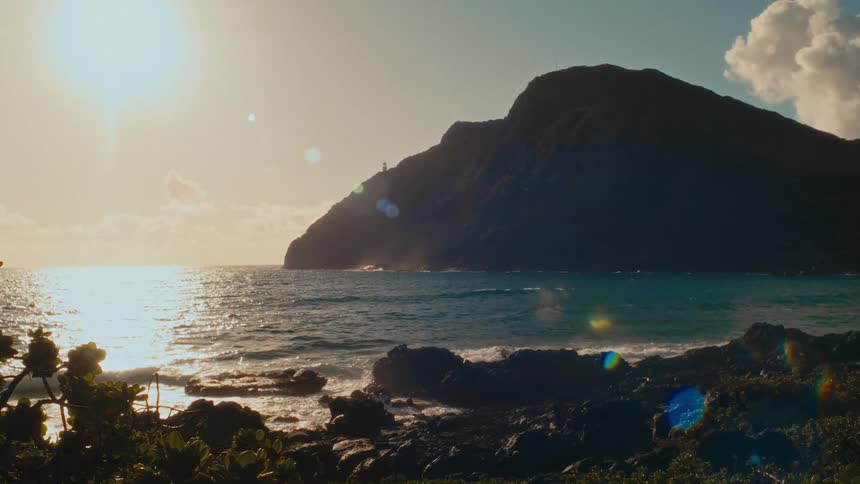The sun shines brightly over a tranquil ocean as waves crash in slow motion against a rocky shore next to a silhouetted mountain