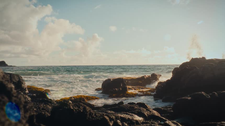 Ocean waves crash in slow motion against a rocky, tropical shore during a bright, sunny morning