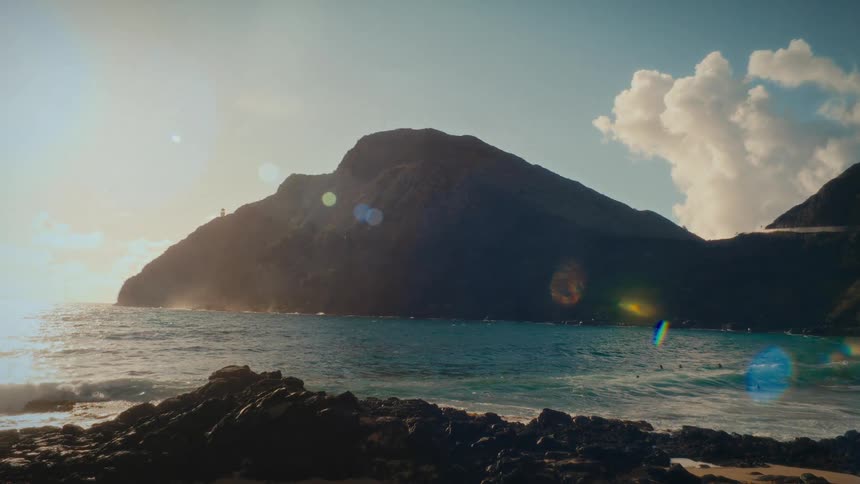 A stunning camera pan across a Hawaiian beach cove reveals a brilliant sunrise over the majestic ocean and mountains