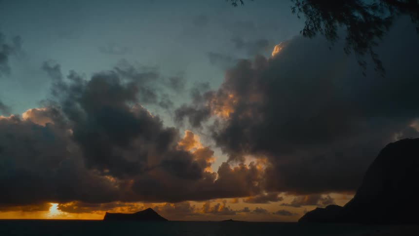 A time-lapse captures a dramatic sunrise over a tropical ocean, with the sun's golden light breaking through and illuminating dark, heavy clouds