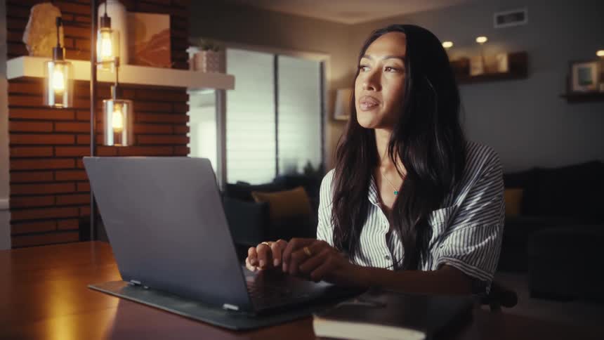 A focused Asian woman works from home on her laptop computer, pausing to think while sitting at a table in her modern living space