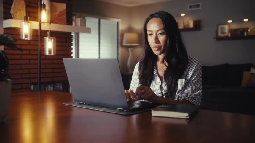 A focused woman works from home on her laptop at a dining room table in a warmly lit house