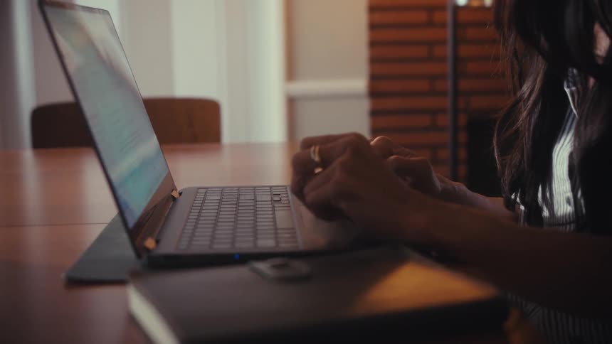 A focused businesswoman works on her laptop at home, with the camera tilting down from her face to her hands as she types