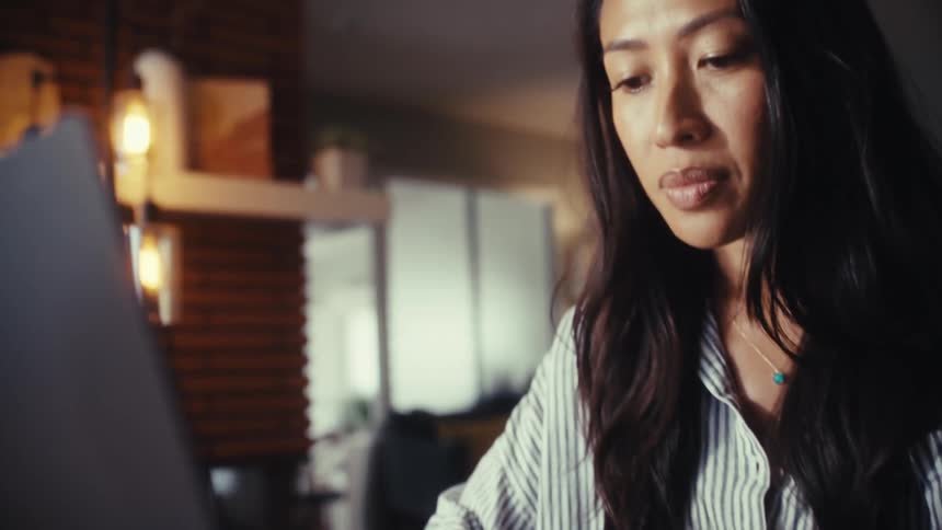 A focused Asian woman types on her laptop in a warmly lit modern home office, concentrating on her work