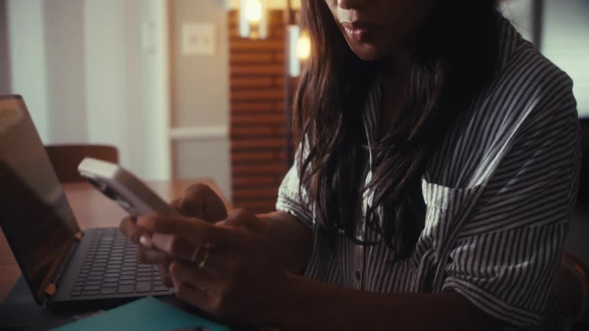 A focused woman works from home at her dining room table, multitasking between her laptop and smartphone