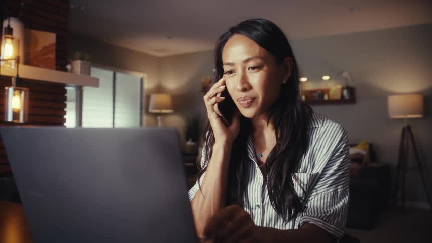 A professional Asian woman works from home in the evening, having a pleasant conversation on her phone while looking at her laptop