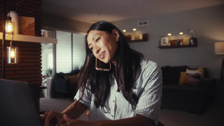 A focused Asian woman multitasks at her dining room table, answering a phone call while continuing to work on her laptop in the evening
