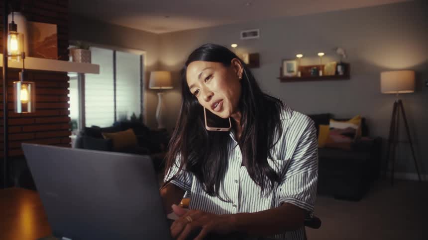 An Asian woman multitasks by taking a phone call while working on her laptop in her warmly lit home in the evening