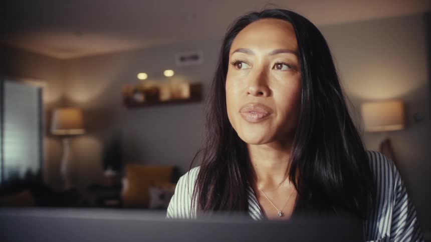 A focused woman works on her laptop in her cozy home at night, pausing to think thoughtfully before continuing