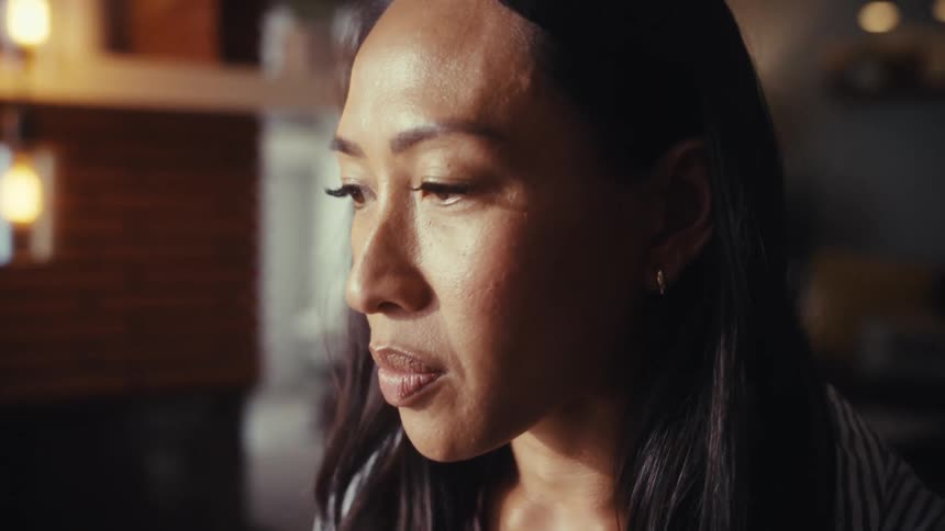 A slow push-in shot from behind reveals a woman working on her laptop at a dining table in a sunlit room, ending in a close-up of her focused face