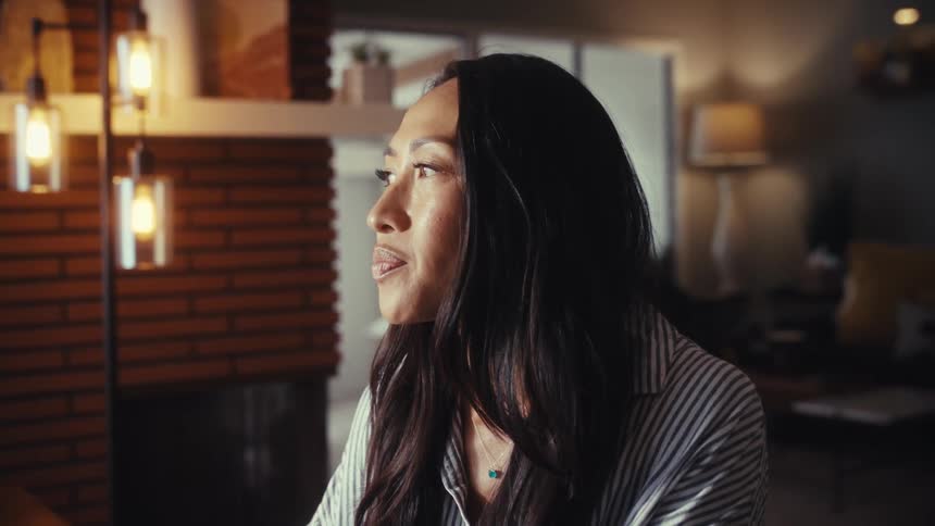 A thoughtful Asian woman working on her laptop in a warmly lit home office breaks into a smile of relief and satisfaction