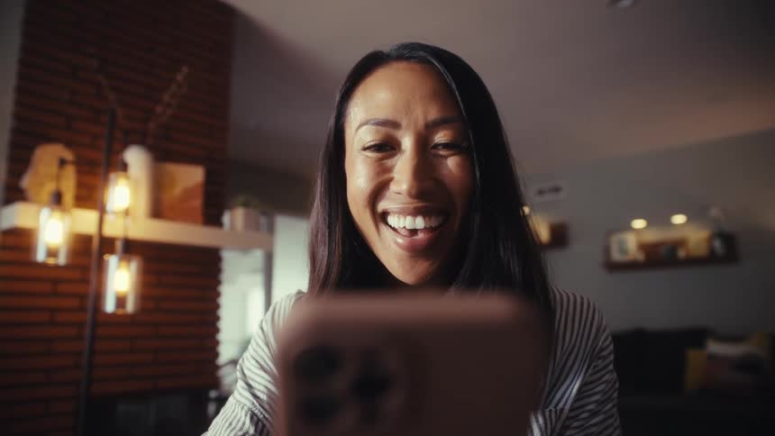 A happy woman reacts with a big smile and laughter while watching something on her mobile phone at home