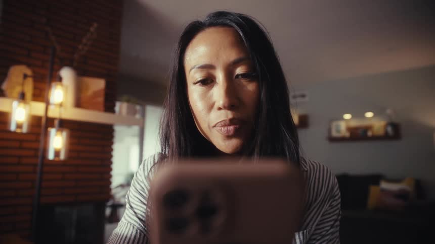 A close-up slow zoom shot of an Asian woman intently focused on her smartphone while working from home