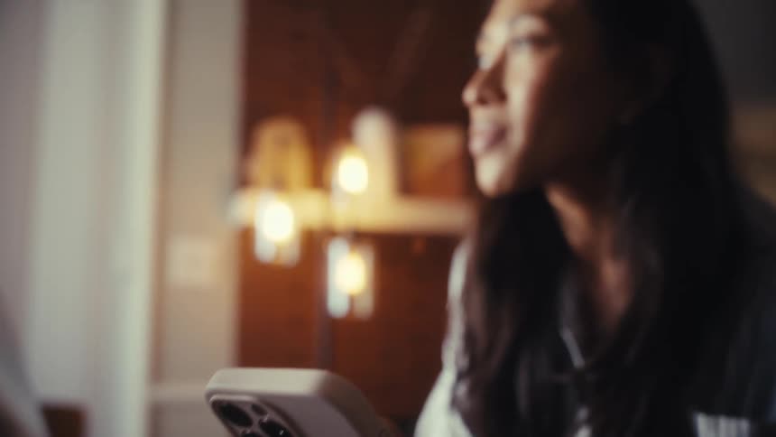 A woman scrolls on her smartphone at a desk with a laptop, the camera slowly revealing her focused expression in a warm, cozy setting