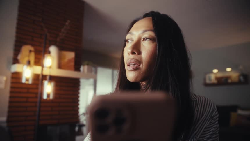 A close-up of a thoughtful Asian woman pausing while looking at her smartphone in a dimly lit room