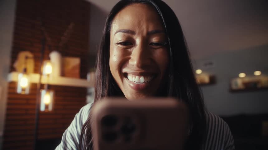 A woman's face lights up with a joyful smile and laughter as she looks at her smartphone in a cozy room