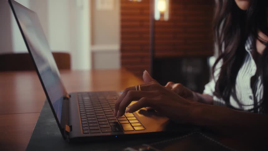 Side-view close-up of a woman's hands typing on a laptop keyboard in a warm, cozy setting