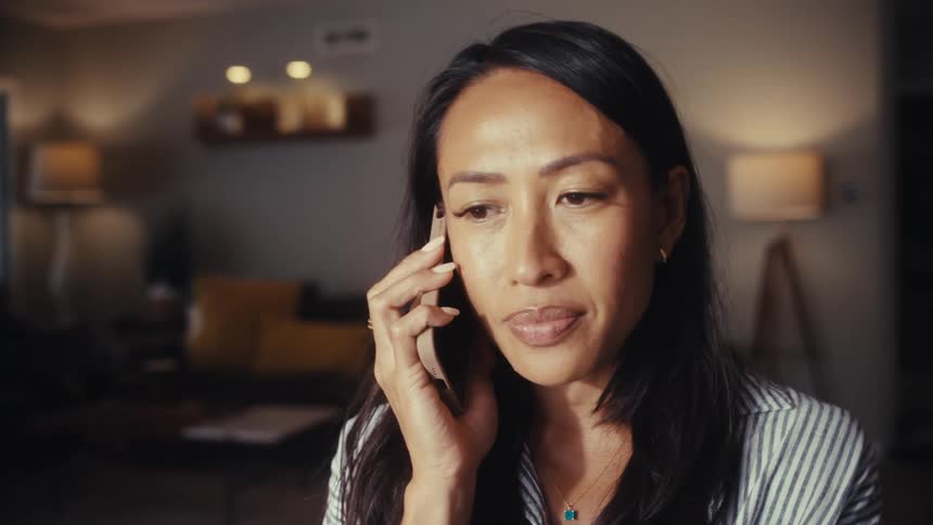 A pensive woman listens intently to a serious phone call while at home in the evening