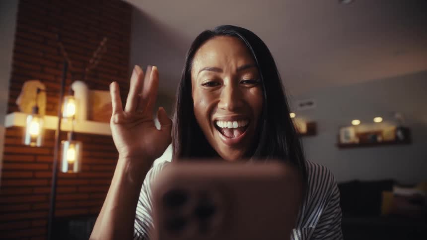 A happy woman smiles and waves while on a video call on her smartphone in her cozy living room