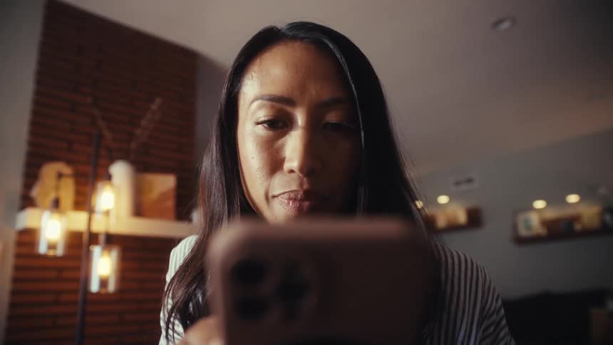 A vertical, low-angle shot reveals a woman looking down intently at her smartphone while sitting at her desk at home