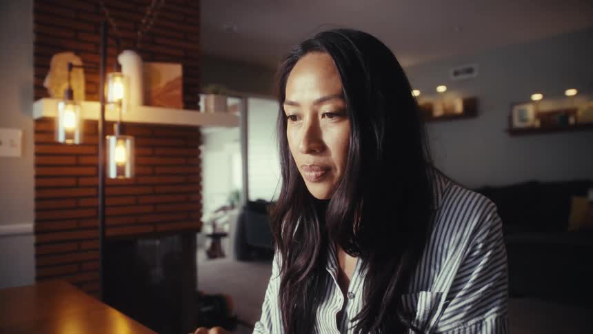 A focused Asian woman types on her laptop while working from her cozy and modern home, with the camera slowly zooming in on her concentrated expression