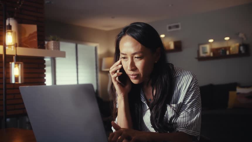 Slow push-in on a serious woman multitasking on her laptop while on a business call from her home in the evening