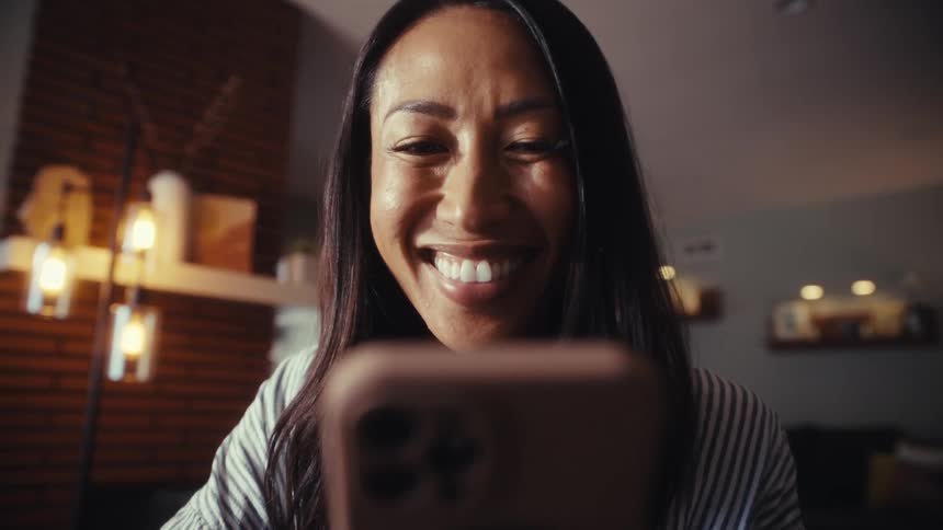 A woman's face, illuminated by her phone, breaks into a wide smile as she reads something in her living room