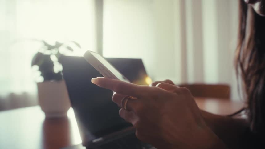 Cinematic slow-motion shot of a woman using her smartphone at a desk with a laptop nearby