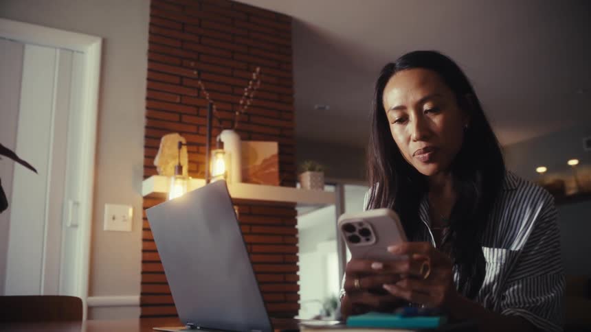 A woman works from home at her dining table, intently scrolling on her smartphone in a softly lit room