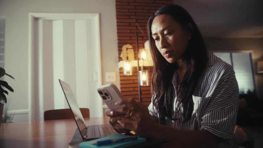 A woman scrolls through her smartphone while working from home at her dining room table with her laptop open