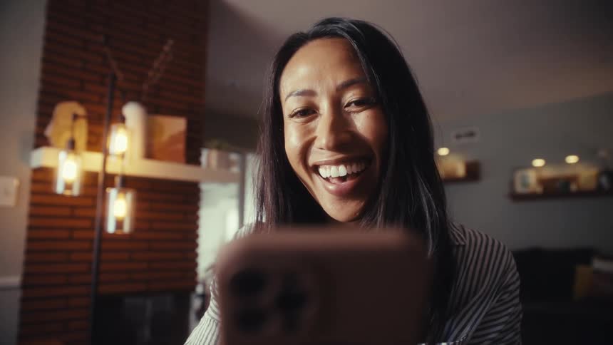 A close-up slow-motion shot of an Asian woman's face as she reacts with a huge smile and laughter to something on her smartphone