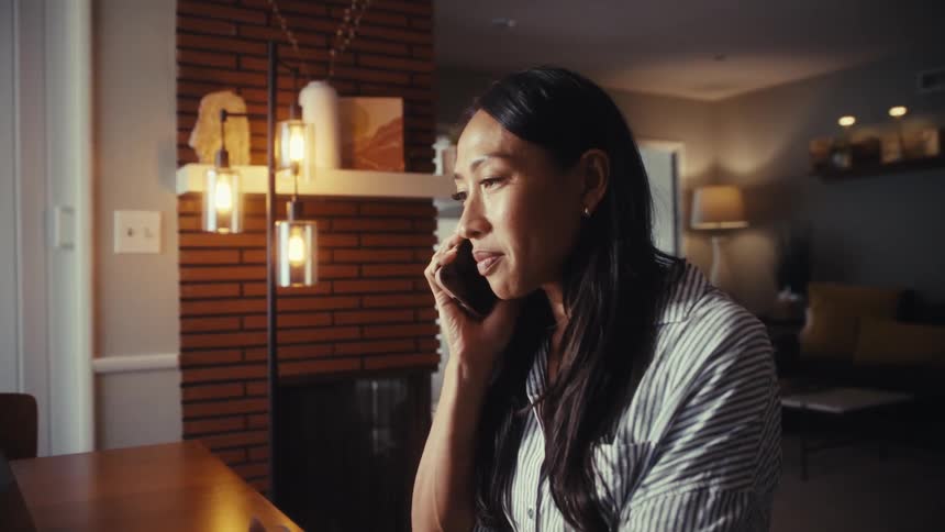 An Asian woman working from home on her laptop at the dining table takes a phone call and smiles while talking