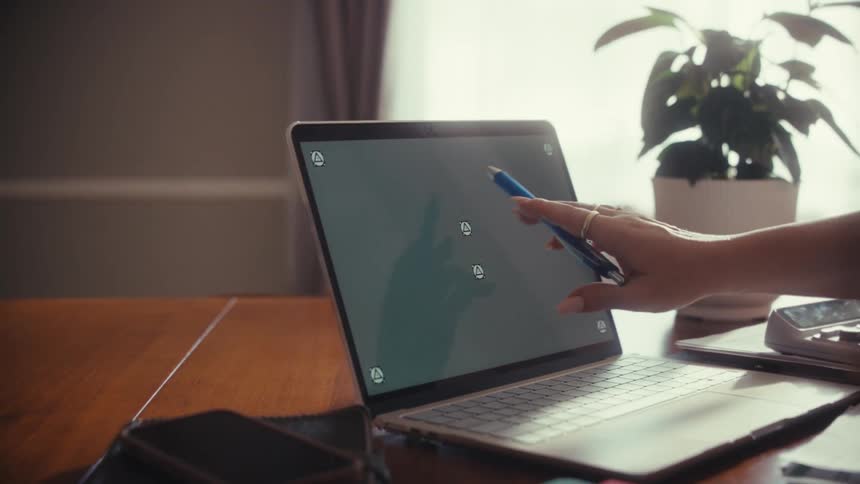 Over-the-shoulder shot of a person using a pen to point at a laptop with a green screen on a wooden desk in a home office