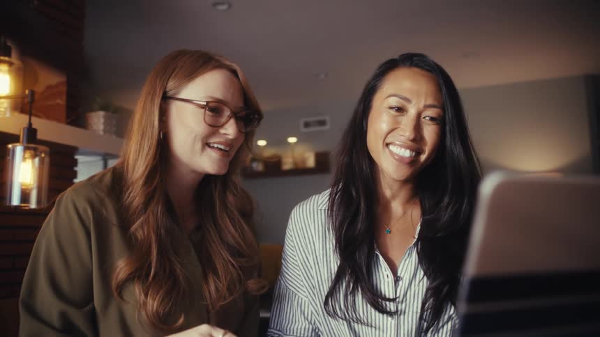 Two diverse female colleagues laugh and wave excitedly during a video conference call on a laptop in a cozy home office