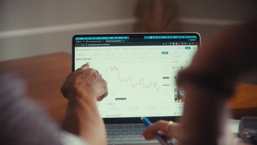 Over-the-shoulder shot of two people analyzing a fluctuating cryptocurrency price chart on a laptop screen