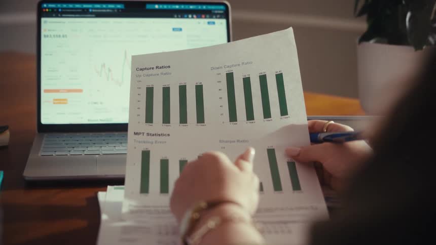 An over-the-shoulder view of a woman analyzing financial performance data on a printed chart with a laptop showing stock market graphs in the background