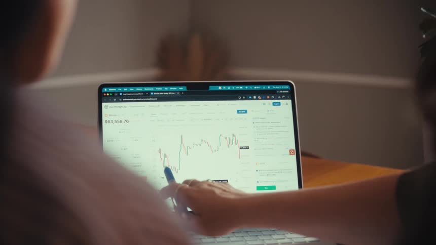 A woman uses a pen to point out trends on a cryptocurrency chart displayed on a laptop to a colleague