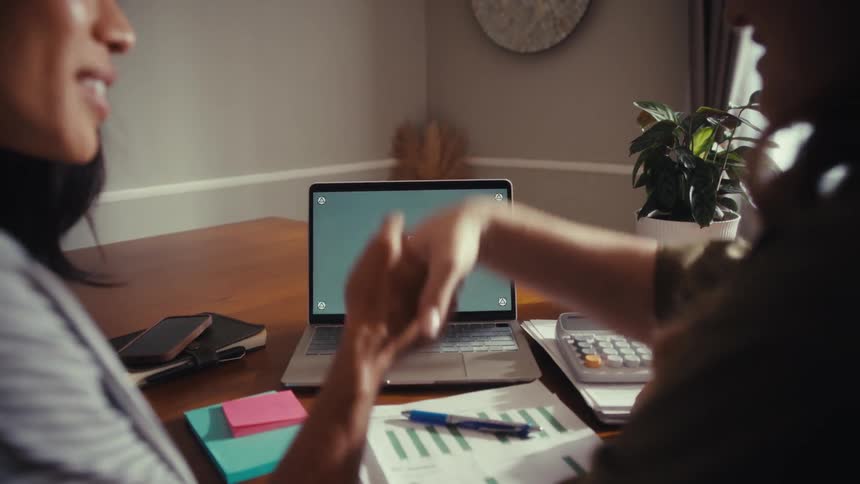 Two diverse businesswomen collaborate on a project at a desk, pointing at a green screen laptop before shaking hands in agreement