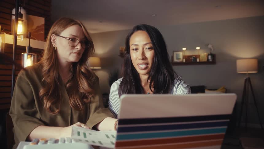Two diverse women collaborate on a project, analyzing charts and data on a laptop in a cozy home office