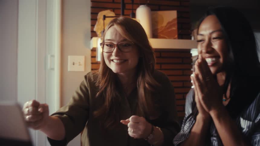 Two diverse businesswomen get excited and celebrate a huge success while looking at a laptop in a stylish home office