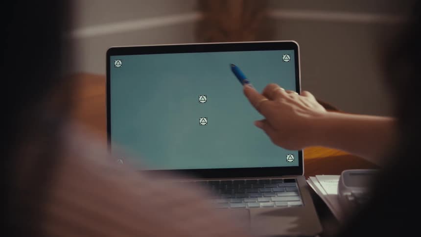 An over-the-shoulder close-up of a person pointing with a pen at a laptop screen with tracking markers during a collaborative meeting