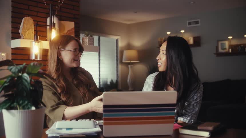 Two diverse female entrepreneurs happily shake hands in their home office after successfully completing a project on their laptop